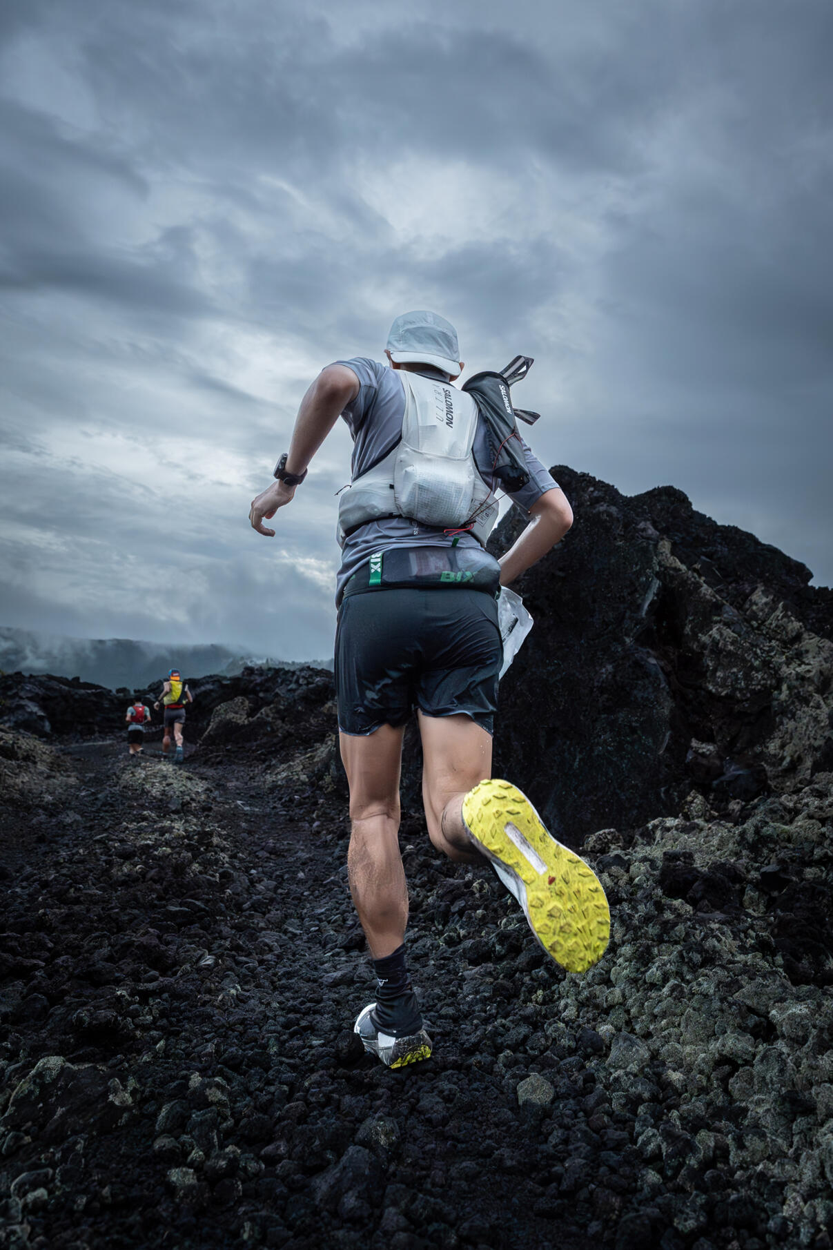 Trail runner ascending rocky volcanic terrain wearing hydration pack, dramatic stormy sky overhead, yellow trail markers visible