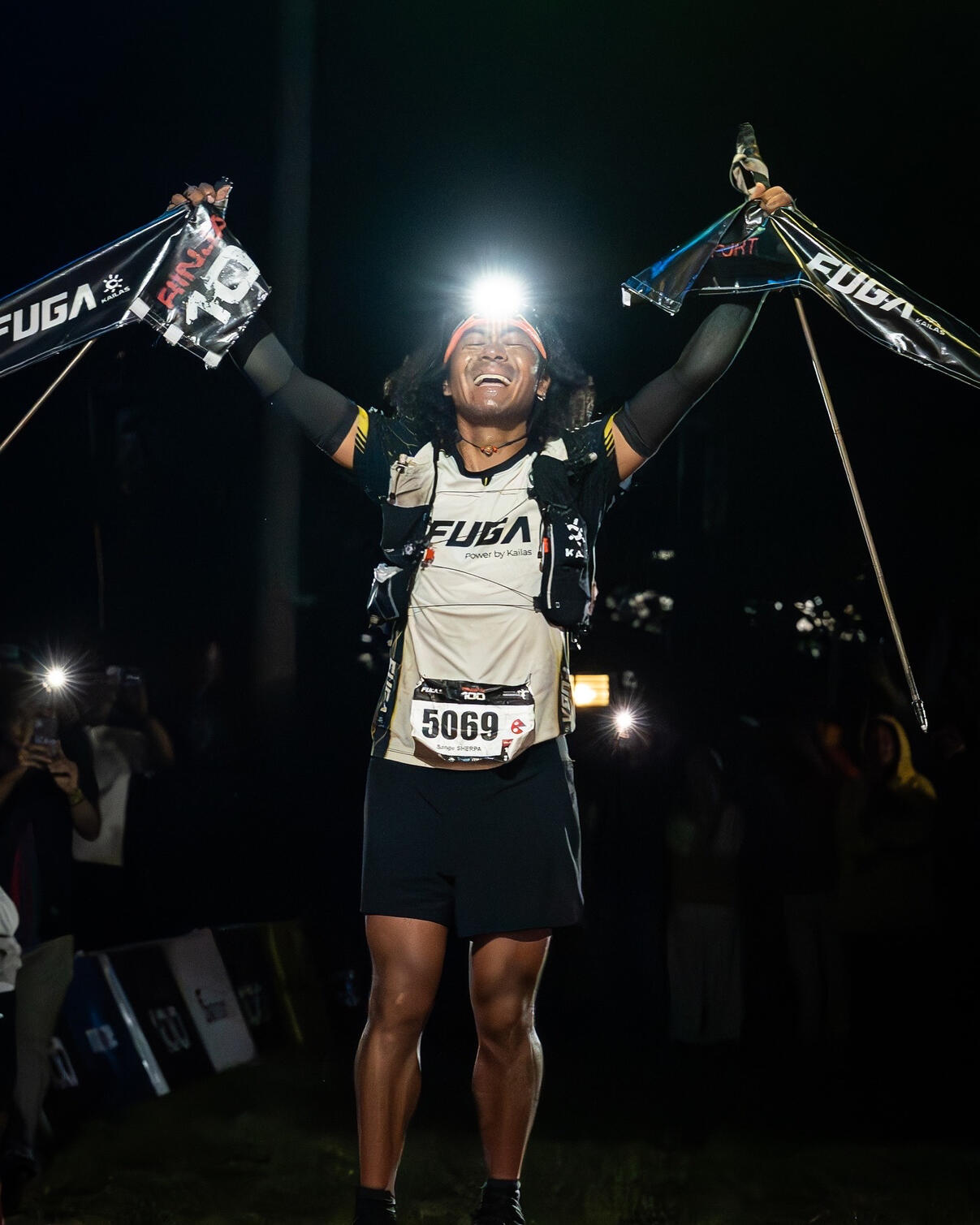 Sange Sherpa Rinjani Ultra Night ultra marathon runner with race bib 5069 and headlamp raising arms in celebration at finish line, wearing hydration vest against dark background