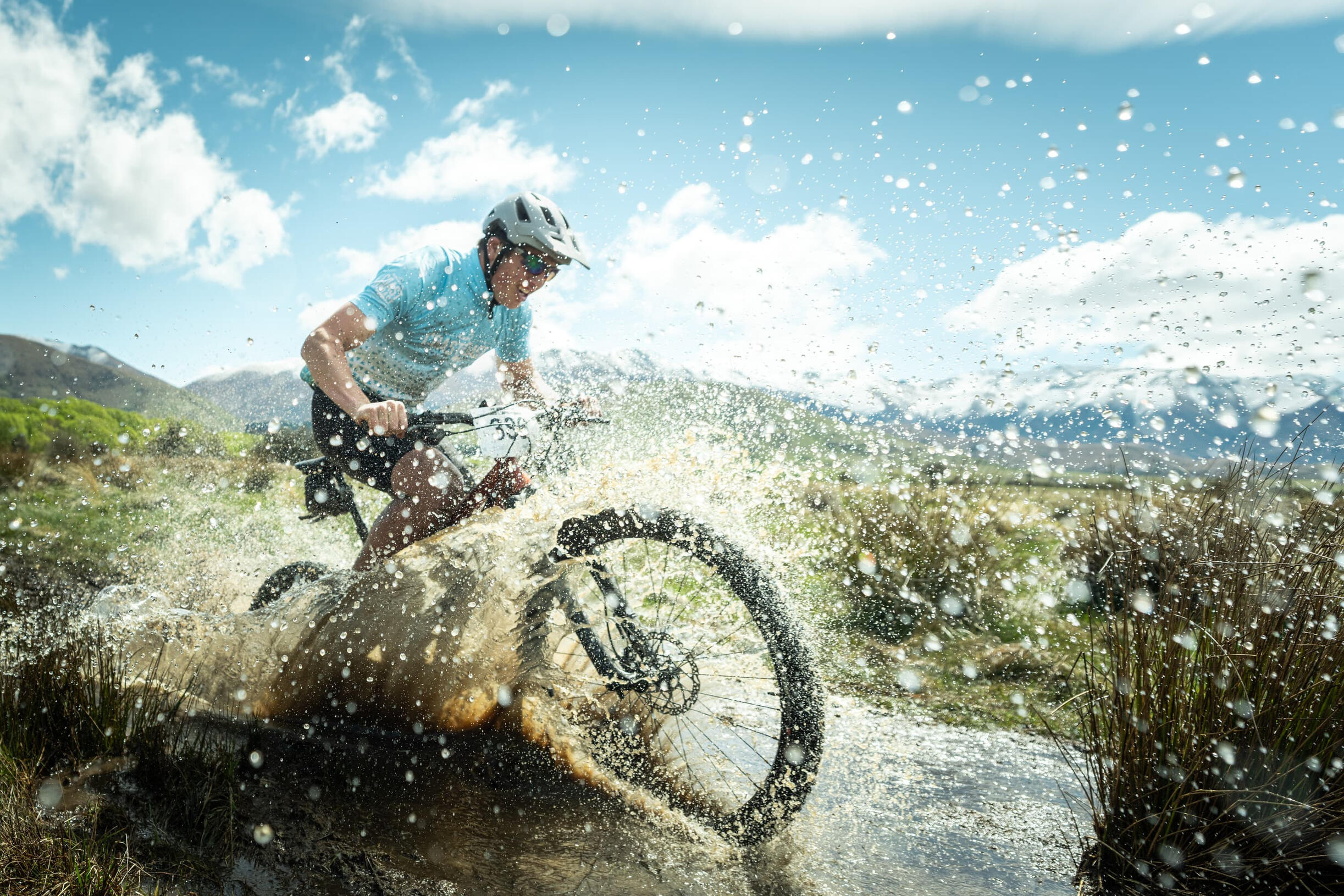 Meridian Twizel Hard Labour Mountain biker riding through water puddle creating massive splash, dramatic snow-covered peaks visible behind in New Zealand alpine setting
