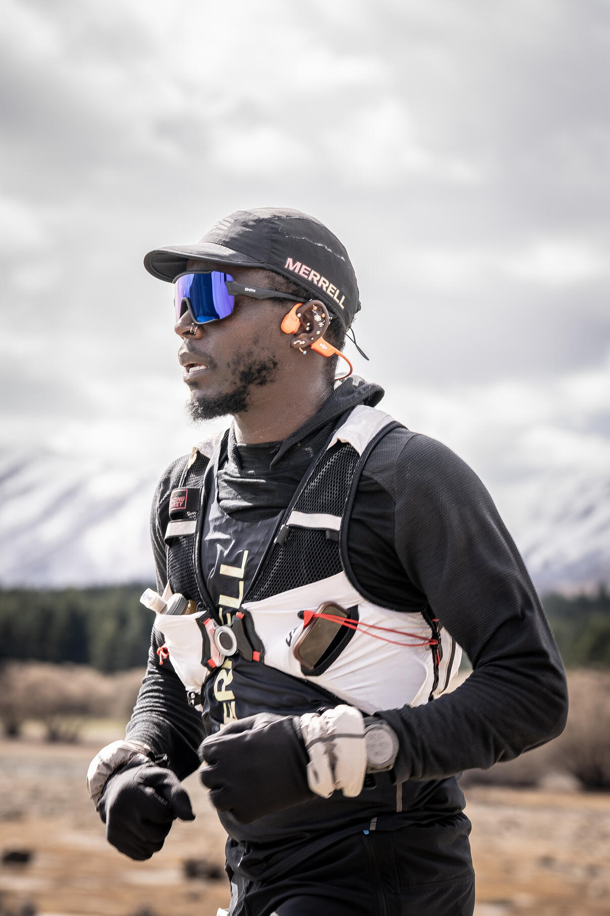 Coree Woltering Athlete Race Tekapo Male ultra runner in black gear with reflective sunglasses and hydration pack running on rocky mountain terrain under cloudy sky