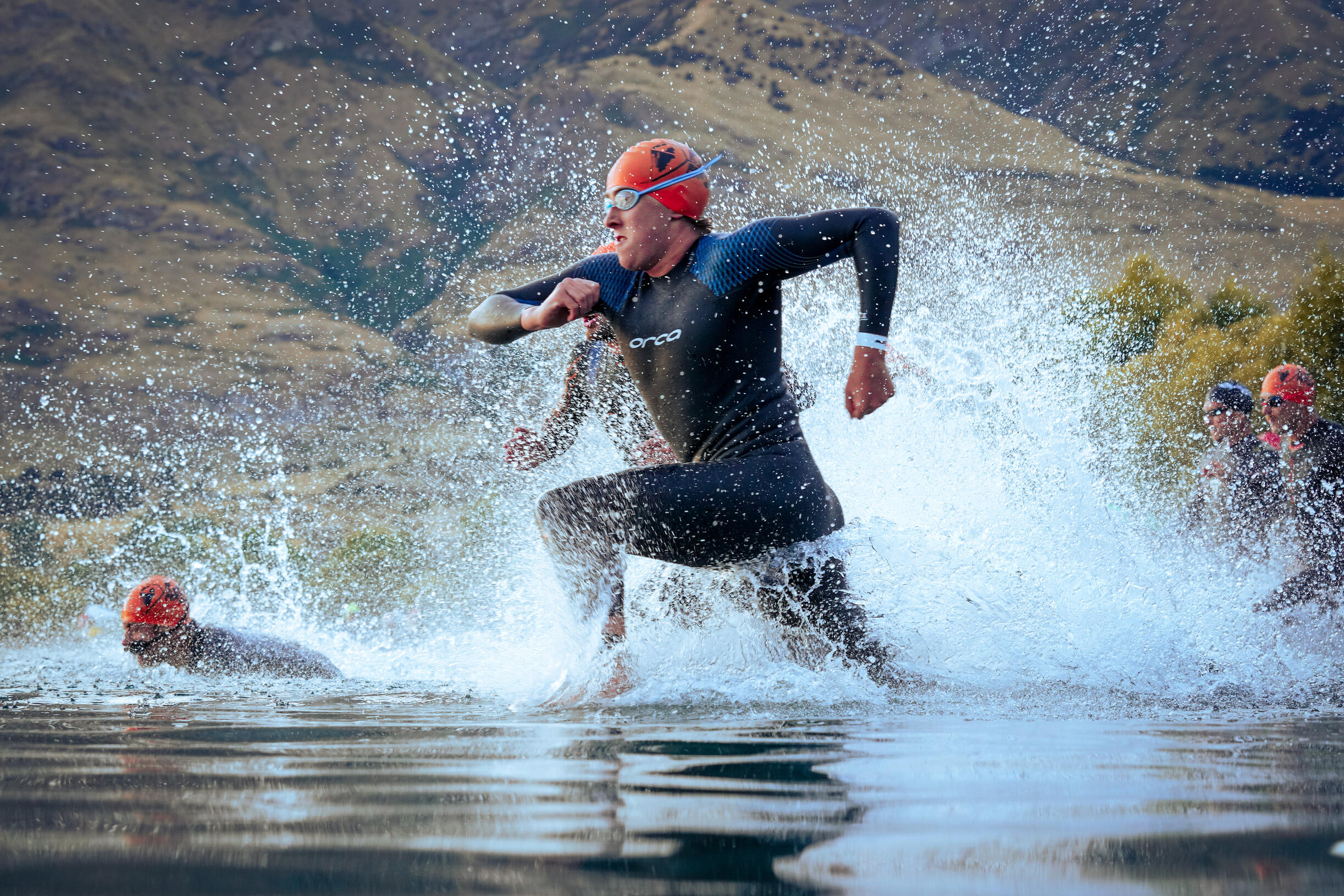 Challenge Wanaka Triathlete in wetsuit and red swim cap exploding out of lake water at race start, mountains visible in background, dramatic splash and motion