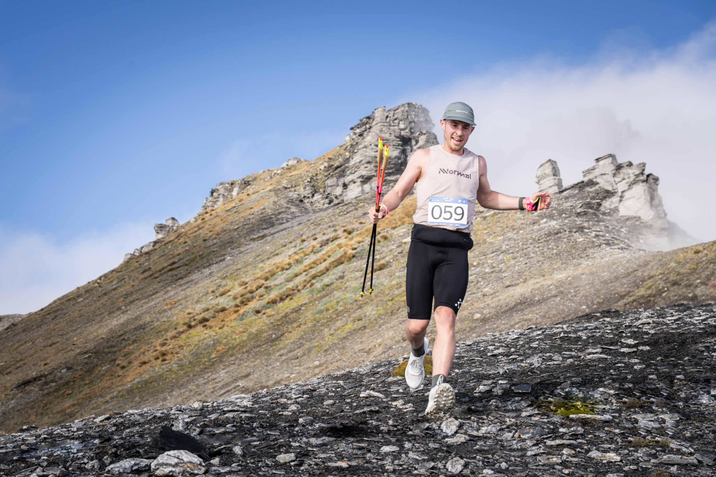 Luke Wilson Treble Cone Mountain runner with race bib 059 ascending volcanic scree slope with trekking pole, dramatic rocky peak and blue sky behind